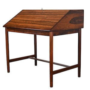 May include: A vintage writing desk with a slanted top and a warm, dark wood grain finish. The desk has a rectangular shape with four slender legs and a horizontal support bar. A small metal ring is attached to the top.