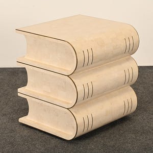 May include: A light beige side table shaped like a stack of three books. The table features a smooth, light-colored surface with dark, thin, vertical lines on the curved edges that mimic the appearance of book pages and spine details.