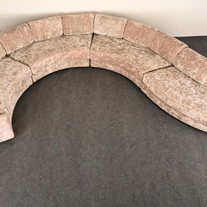 May include: A curved, sectional sofa with a light pink, textured fabric. The sofa is composed of multiple sections, forming a semi-circular shape. The design is modern and the color is neutral, suitable for various interior styles.