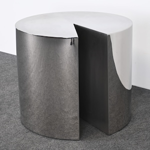 May include: A modern, cylindrical side table with a unique pie-slice cutout. The table is made of a brushed metal material, with a silver-gray finish. The top surface is a lighter shade of silver. The table is approximately 20 inches in diameter and 18 inches tall.