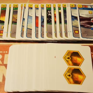 Può includere: Una collezione di carte da gioco rettangolari disposte su due file. La fila superiore mostra carte con illustrazioni colorate, valori numerici in esagoni gialli e testo. La fila inferiore presenta carte bianche con due esagoni gialli sovrapposti contenenti immagini.