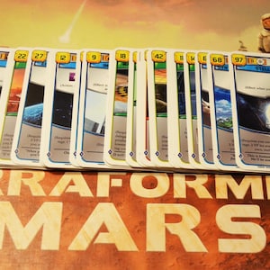 Könnte beinhalten: Eine Sammlung von Spielkarten aus dem Brettspiel "Terraforming Mars". Die Karten haben einen weißen Rand mit blauen Akzenten und zeigen verschiedene Bilder und Text. Der Spieltitel ist unten im Bild sichtbar.