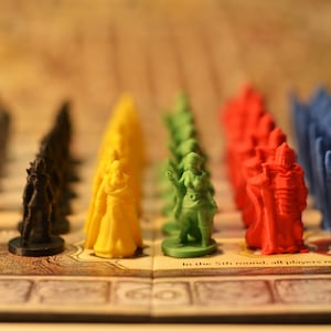 Puede incluir: Primer plano de un juego de mesa con piezas de juego de plástico de colores. Las piezas están dispuestas en filas de diferentes colores, incluyendo gris, negro, amarillo, verde, rojo y azul. Las piezas del juego están en un tablero con un fondo marrón y una etiqueta de texto blanco que dice "En la 5ª ronda, todos los jugadores reciben 1 adicional".