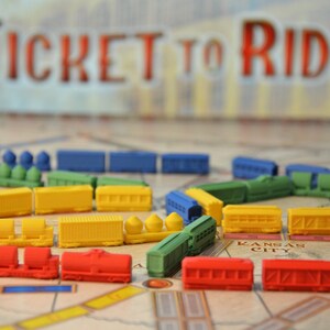 Peut inclure: Gros plan sur un plateau de jeu avec des pièces de train colorées. Le plateau de jeu présente une carte des États-Unis avec des villes et des itinéraires. Les pièces de train sont de différentes couleurs, notamment rouge, bleu, vert et jaune. Le jeu s'appelle "Ticket to Ride".