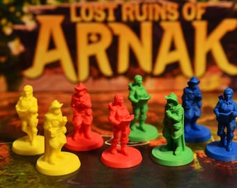 May include: A collection of eight miniature plastic explorer figurines in yellow, red, green, and blue, each standing on a circular base. The figures are positioned in front of a board game box featuring the text Lost Ruins of Arnak.