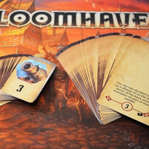Peut inclure: Gros plan sur deux piles de cartes du jeu de société Gloomhaven. La carte du dessus de la pile de gauche montre un canon sur fond bleu avec le numéro 3. La carte du dessus de la pile de droite contient le texte suivant : "La ville a décidé de mettre en place une exposition sur la faune locale. Ramenez quelques exemples pour faire partie de leur exposition." et le numéro 3.