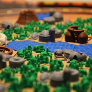 以下が含まれることがあります： 3Dプラスチック製の駒を使用したモジュール式ヘキサゴンマップのボードゲーム。ボードには、緑色の森の集まり、青いテクスチャの水タイル、茶色の山岳地帯、そして様々な灰色と白色の建造物が配置されています。