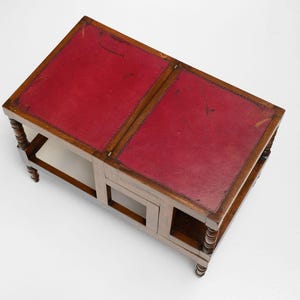 Puede incluir: Una mesa de centro de madera con tapa de cuero rojo. La mesa tiene forma rectangular con dos secciones y un estante inferior. Las patas son torneadas y la mesa es de madera.