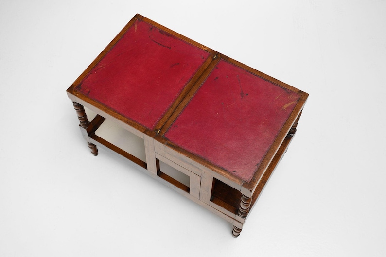 Puede incluir: Una mesa de centro de madera con tapa de cuero rojo. La mesa tiene forma rectangular con dos secciones y un estante inferior. Las patas son torneadas y la mesa es de madera.