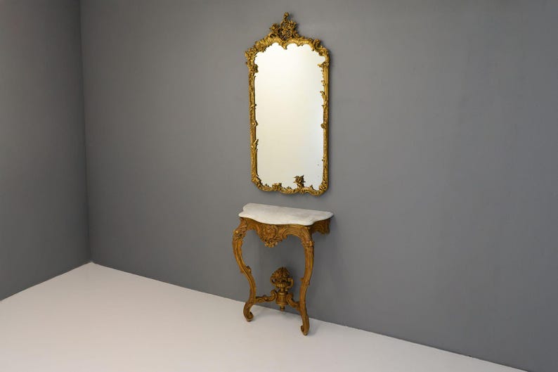 Peut inclure: Un miroir dor&eacute; avec des d&eacute;tails orn&eacute;s au-dessus d'une console peinte en or avec un plateau en marbre blanc.