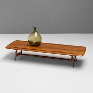 May include: A mid-century modern style coffee table with a long, rectangular wooden top and a lower shelf. A large, green glass bottle sits on the table. The table has a warm, natural wood finish and is supported by angled legs.