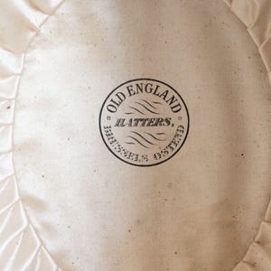 May include: Close-up of the interior of a hat, featuring a cream-colored satin lining with a ruffled edge. A circular stamp in black ink reads "OLD ENGLAND HATTERS, BRUSSELS OSTEND."