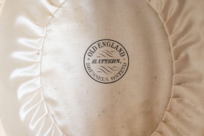 May include: Close-up of the interior of a hat, featuring a cream-colored satin lining with a ruffled edge. A circular stamp in black ink reads "OLD ENGLAND HATTERS, BRUSSELS OSTEND."