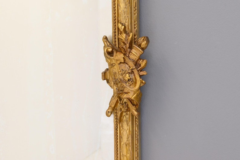 May include: A gold-colored ornate frame with intricate carvings and a decorative design. The frame is made of wood and has a smooth, polished finish.