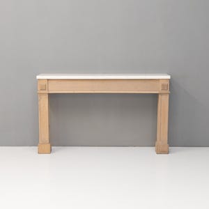 May include: A light-colored wooden fireplace mantel with a white marble top. The mantel has a rectangular design with two square columns on each side. The background is a solid gray wall and a white floor.