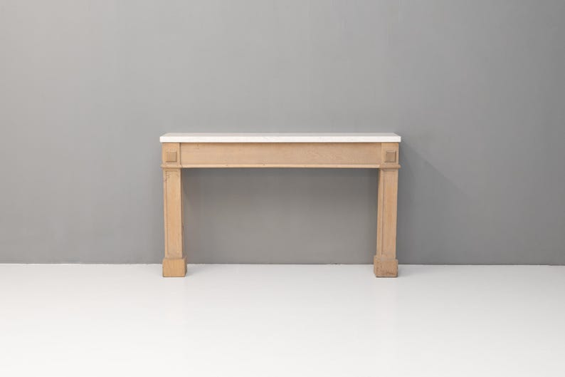 May include: A light-coloured wooden fireplace mantel with a white marble top. The mantel has a rectangular design with two square columns on each side. The background is a solid grey wall and a white floor.