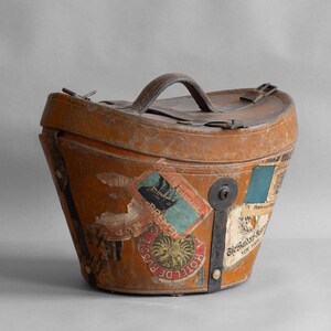 May include: Vintage brown leather travel trunk with a handle and metal hardware. The trunk is adorned with colorful travel stickers, including one that says "Hotel de Russie". The trunk has a keyhole and a leather strap.