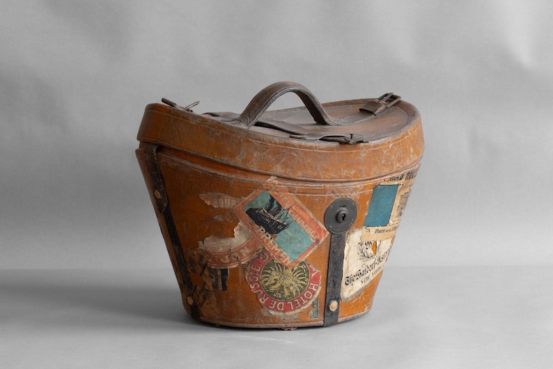 May include: Vintage brown leather travel trunk with a handle and metal hardware. The trunk is adorned with colorful travel stickers, including one that says "Hotel de Russie". The trunk has a keyhole and a leather strap.