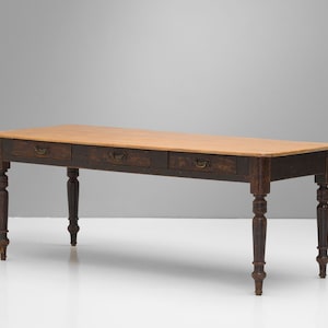 Può includere: Un lungo tavolo in legno scuro con tre cassetti e gambe tornite. Il piano del tavolo è di un colore di legno più chiaro.