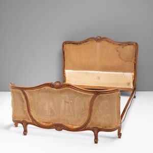 May include: A wooden bed frame featuring a headboard and footboard with curved, carved edges and shell motifs. The central panels are upholstered in a light brown, textured fabric. The frame is supported by short, carved wooden legs.