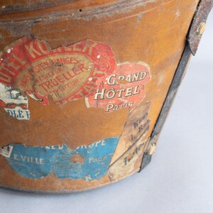 May include: Close-up of a vintage, weathered leather bucket with travel stickers. The stickers feature text like "Grand Hotel Paris" and "Hotel Kor". The bucket has metal accents and a worn appearance.
