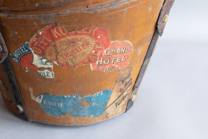 May include: Close-up of a vintage, weathered leather bucket with travel stickers. The stickers feature text like "Grand Hotel Paris" and "Hotel Kor". The bucket has metal accents and a worn appearance.