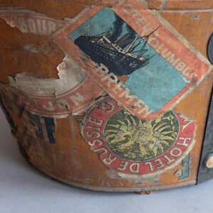 May include: Close-up of a vintage travel trunk with several aged stickers. The stickers feature images of a ship, a hotel emblem, and other travel-related graphics. The trunk is made of brown leather and has metal accents.