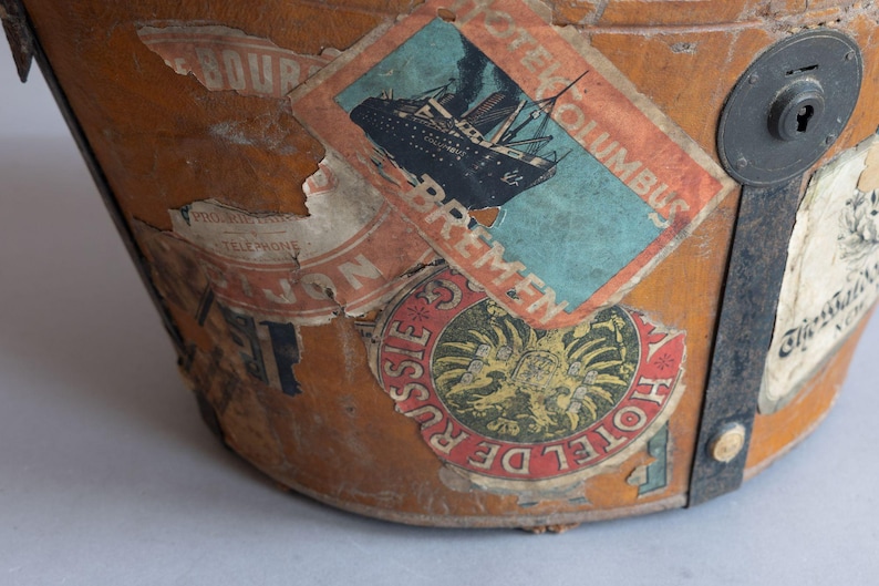 May include: Close-up of a vintage travel trunk with several aged stickers. The stickers feature images of a ship, a hotel emblem, and other travel-related graphics. The trunk is made of brown leather and has metal accents.