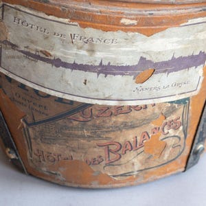 May include: Close-up of a vintage, cylindrical travel trunk with peeling labels. The trunk is a warm, aged brown with black straps and metal accents. Labels include the text "HOTEL DE FRANCE" and "HOTEL des BALANCES".