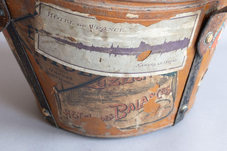May include: Close-up of a vintage, cylindrical travel trunk with peeling labels. The trunk is a warm, aged brown with black straps and metal accents. Labels include the text "HOTEL DE FRANCE" and "HOTEL des BALANCES".