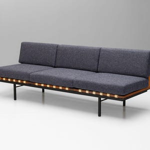 Op de afbeelding: Een driezitsbank met een donkergrijze stoffen bekleding en een houten frame. De bank heeft een zwarte metalen basis met een latten ontwerp.