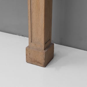 May include: A section of a light brown, rectangular wooden pillar. The pillar has a square base and a slightly textured surface. The background is a light grey wall and a white floor.