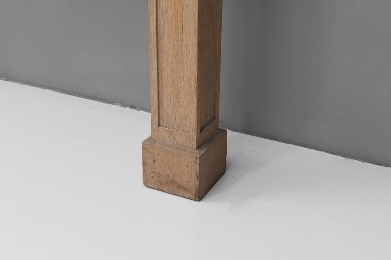May include: A section of a light brown, rectangular wooden pillar. The pillar has a square base and a slightly textured surface. The background is a light grey wall and a white floor.