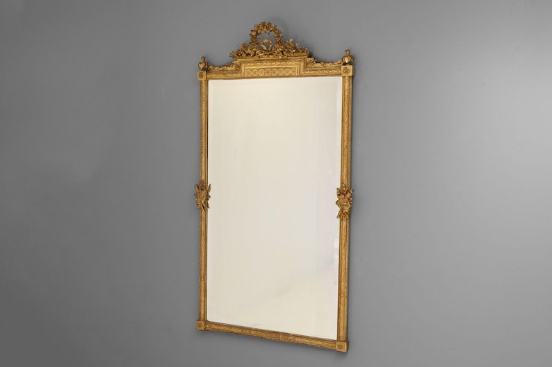 May include: A large, ornate gold mirror with a decorative frame. The frame is carved with intricate details and has a central crest at the top.