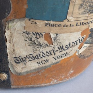May include: Close-up of a vintage, weathered leather hat with various aged labels. The labels feature text including "The Waldorf-Astoria New York" and "Place de la Libert&eacute; et des Palm". The hat has metal hardware and a keyhole.