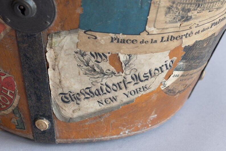 May include: Close-up of a vintage, weathered leather hat with various aged labels. The labels feature text including "The Waldorf-Astoria New York" and "Place de la Libert&eacute; et des Palm". The hat has metal hardware and a keyhole.