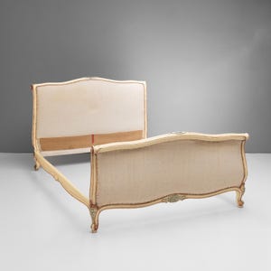 May include: A bed frame featuring a cream-colored wooden structure with curved headboard and footboard panels upholstered in a light beige fabric. The frame includes decorative carved details at the center and legs, with visible nailhead trim.
