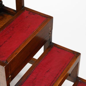 Puede incluir: Un taburete de biblioteca de madera con tres escalones. Cada escal&oacute;n tiene una incrustaci&oacute;n de cuero rojo. La madera es de color marr&oacute;n c&aacute;lido y el cuero tiene un aspecto envejecido. El taburete est&aacute; dise&ntilde;ado para alcanzar estantes altos.