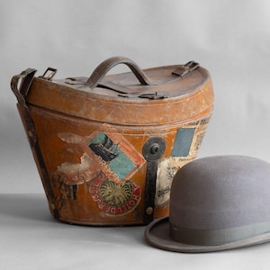 May include: An antique, weathered leather travel case with a top handle and various travel stickers. A gray bowler hat sits beside the case. The case and hat evoke a vintage travel theme.