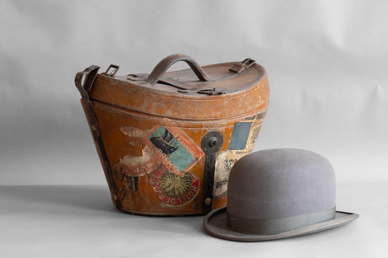 May include: An antique, weathered leather travel case with a top handle and various travel stickers. A gray bowler hat sits beside the case. The case and hat evoke a vintage travel theme.
