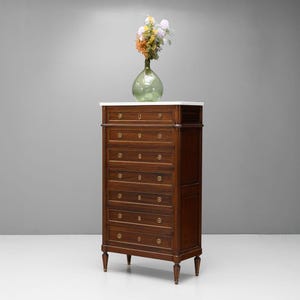 May include: A dark wood chest of drawers with a white marble top. The chest has seven drawers with brass handles. A clear glass vase with flowers sits on top. The chest is a classic piece of furniture, approximately 170 cm tall.