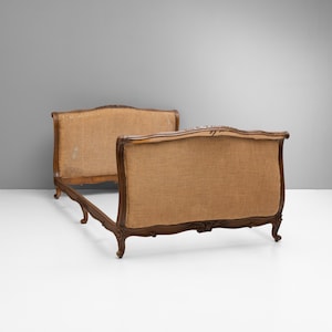 May include: A wooden bed frame featuring a headboard and footboard upholstered in natural tan hessian. The dark brown wood frame displays carved floral details along the top and bottom edges, supported by four curved legs with scroll feet.
