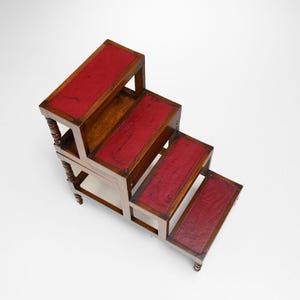 Puede incluir: Un taburete de madera con cuatro escalones, cada uno con una parte superior de cuero rojo. El taburete tiene un acabado marr&oacute;n oscuro con patas torneadas decorativas y un dise&ntilde;o cl&aacute;sico. Los escalones miden unos 30 cm de ancho.
