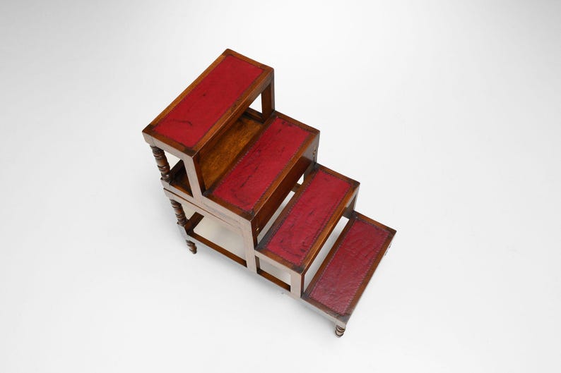 Puede incluir: Un taburete de madera con cuatro escalones, cada uno con una parte superior de cuero rojo. El taburete tiene un acabado marr&oacute;n oscuro con patas torneadas decorativas y un dise&ntilde;o cl&aacute;sico. Los escalones miden unos 30 cm de ancho.