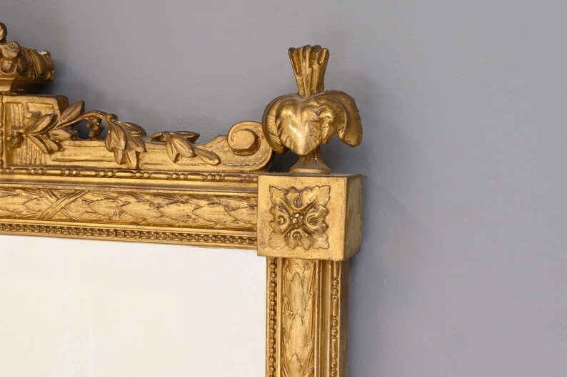 May include: A gold-toned ornate mirror frame with intricate carvings and a decorative top. The frame is made of wood and has a distressed finish.