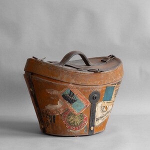 May include: A vintage, tan leather travel hat box with a curved shape and a top handle. The box is adorned with several travel stickers, including one that says "Hotel de Russie". The box has metal hardware and a keyhole.