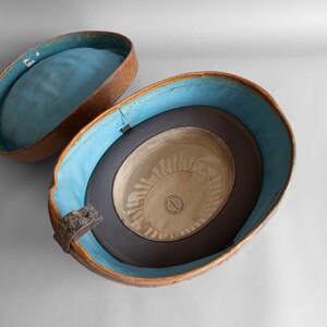 May include: An open, antique hat box with a brown exterior and a light blue interior. Inside the box is a dark gray hat with a cream-colored interior. The hat box is oval-shaped and has a hinged lid.