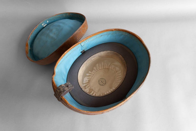 May include: An open, antique hat box with a brown exterior and a light blue interior. Inside the box is a dark gray hat with a cream-colored interior. The hat box is oval-shaped and has a hinged lid.