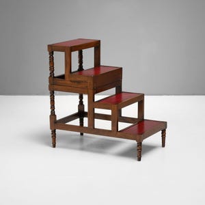 Puede incluir: Un taburete de madera de tres niveles con superficies rojas. El taburete marr&oacute;n oscuro tiene patas torneadas decorativas. El dise&ntilde;o es similar a un peque&ntilde;o taburete de biblioteca.