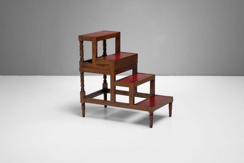 Puede incluir: Un taburete de madera de tres niveles con superficies rojas. El taburete marr&oacute;n oscuro tiene patas torneadas decorativas. El dise&ntilde;o es similar a un peque&ntilde;o taburete de biblioteca.